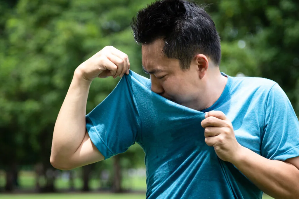 Image d’un homme en t-shirt bleu dans un parc, soulevant son bras pour sentir une odeur corporelle
