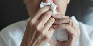 Rhinite Allergique (Rhume des Foins) : Symptômes & Traitement Personne se mouchant dans un mouchoir en papier blanc, symptôme typique de la rhinite allergique ou du rhume des foins, portant un haut blanc dans un environnement intérieur