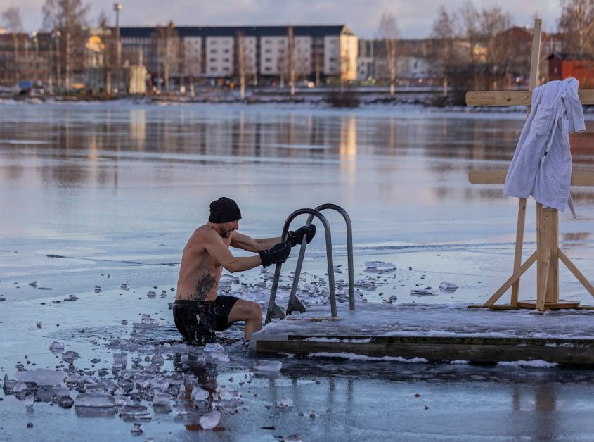 Bénéfices et Risques des Bains de Glace (Thérapie par Eau Froide) Un homme torse nu portant un bonnet noir et un short s'immerge dans un lac gelé à Ludvika, Suède, utilisant une échelle métallique fixée à la glace. Des morceaux de glace brisée flottent autour de lui. Une serviette blanche est accrochée à un support en bois à proximité. En arrière-plan, on aperçoit des immeubles d'habitation et des arbres nus dans la lumière hivernale. La scène illustre la pratique nordique traditionnelle du bain en eau glacée.