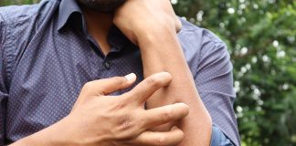 Dermatite Atopique (Eczéma): Symptômes, Causes et Traitement Gros plan sur une personne se grattant le bras à travers une chemise à motifs bleu et rouge. L'image illustre le symptôme de démangeaison caractéristique de la dermatite atopique, prise en extérieur avec un arrière-plan flou de feuillage vert. La photo met en évidence le geste instinctif de grattage associé à l'eczéma, montrant la main droite grattant la partie supérieure du bras gauche.