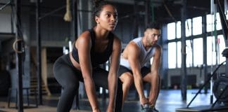 Faire du sport tous les jours : Conseils et sécurité Deux personnes faisant des exercices avec des kettlebells dans une salle de sport moderne, montrant une séance d'entraînement en binôme avec une femme en premier plan portant une tenue de sport noire et un homme en arrière-plan portant un débardeur gris