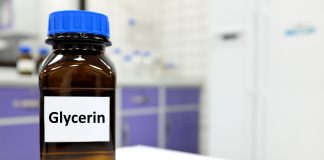 Glycérine : Avantages, Utilisations et Où la Trouver Flacon brun de glycérine avec un bouchon bleu sur une table de laboratoire blanche, avec des armoires violettes floues en arrière-plan, illustrant un ingrédient courant dans les cosmétiques et les produits de soin.