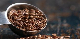 Graines de lin : 8 bienfaits pour la santé et comment les consommer Cuillère à mesurer remplie de graines de lin brunes avec quelques graines dispersées sur une surface texturée sombre