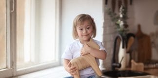 Syndrome de Down : Symptômes & Causes Petite fille assise sur un comptoir de cuisine, tenant un jouet en bois, avec des cartes éducatives, montrant l'apprentissage positif des enfants atteints du syndrome de Down