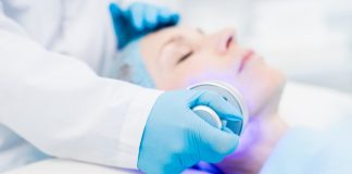 Luminothérapie LED : Fonctionnement, Couleurs, Bienfaits & Risques Traitement de luminothérapie LED en cabinet médical avec praticien portant des gants bleus effectuant un soin du visage sur une patiente allongée sous une lumière LED violette