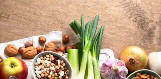 Les 20 Meilleurs Aliments Prébiotiques à Consommer Aliments prébiotiques sains sur une table en bois: poireaux frais, pommes, noix, amandes, ail, oignon, légumineuses mélangées et un verre de lait, illustrant des sources naturelles de fibres pour une bonne santé intestinale