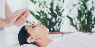 Reiki : Qu’est-ce que c’est, techniques, bienfaits et risques Séance de Reiki dans un cadre zen avec plantes vertes, praticien tenant ses mains au-dessus de la tête d'une cliente allongée sur une serviette blanche
