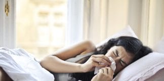 Le Rhume : Symptômes, Rhume vs. Grippe, Traitement Jeune femme alitée avec un rhume, se mouchant dans un mouchoir en papier, allongée dans un lit avec des draps blancs près d'une fenêtre lumineuse, illustrant les symptômes du rhume et de la grippe