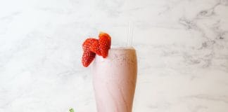12 shakes protéinés sains pour prendre du poids Smoothie rose aux fraises frais et crémeux garni de fraises fraîches, servi dans un grand verre sur fond de marbre blanc