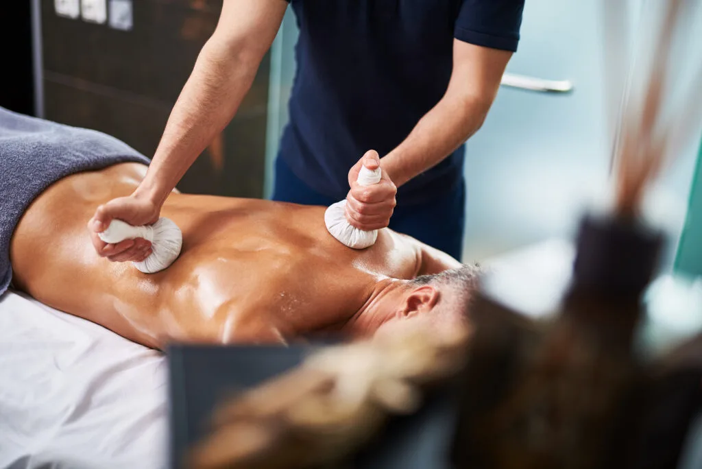 Un thérapeute applique un massage ayurvédique sur le dos d’une personne allongée, utilisant des pochons d’herbes chaudes, dans une pièce calme et relaxante.