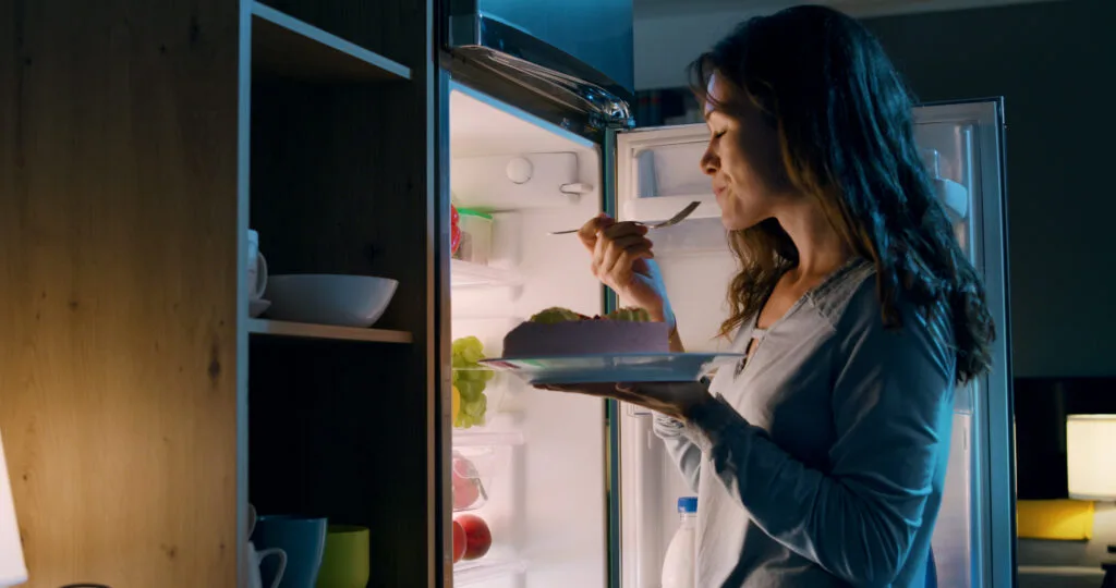 Une femme mange un repas devant un réfrigérateur ouvert la nuit, illustrant les risques de manger juste avant de dormir.