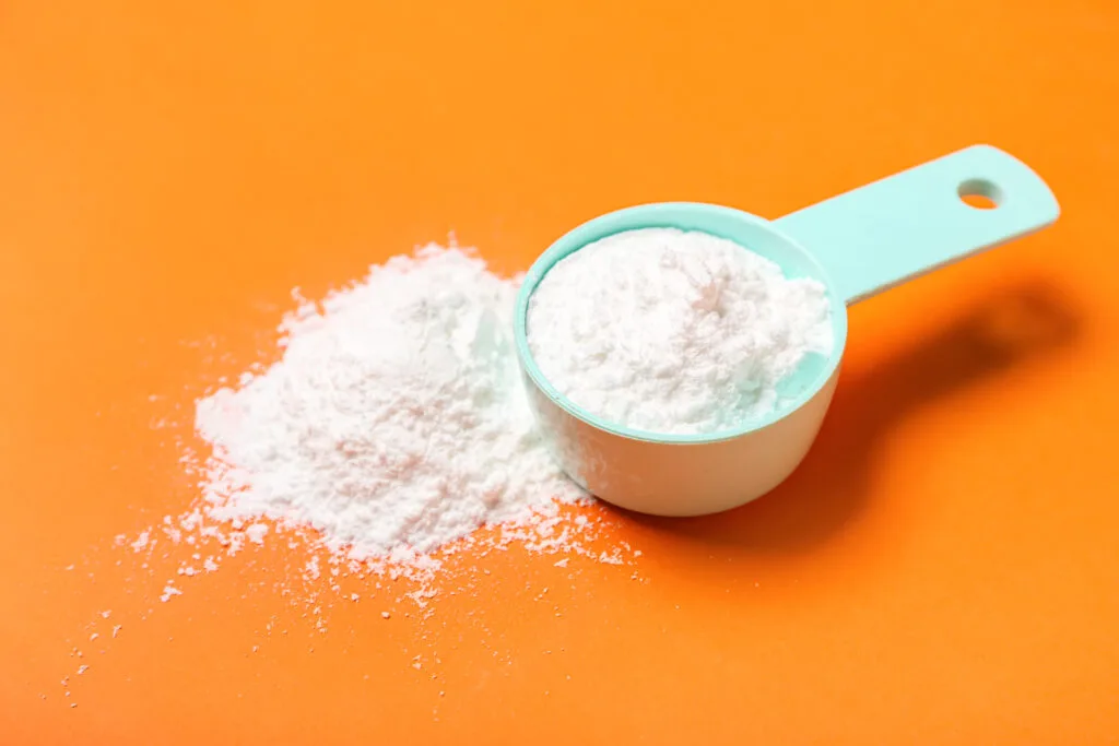 Une cuillère blanche avec un manche bleu contenant de la poudre de créatine monohydrate, partiellement renversée sur une surface orange vif.