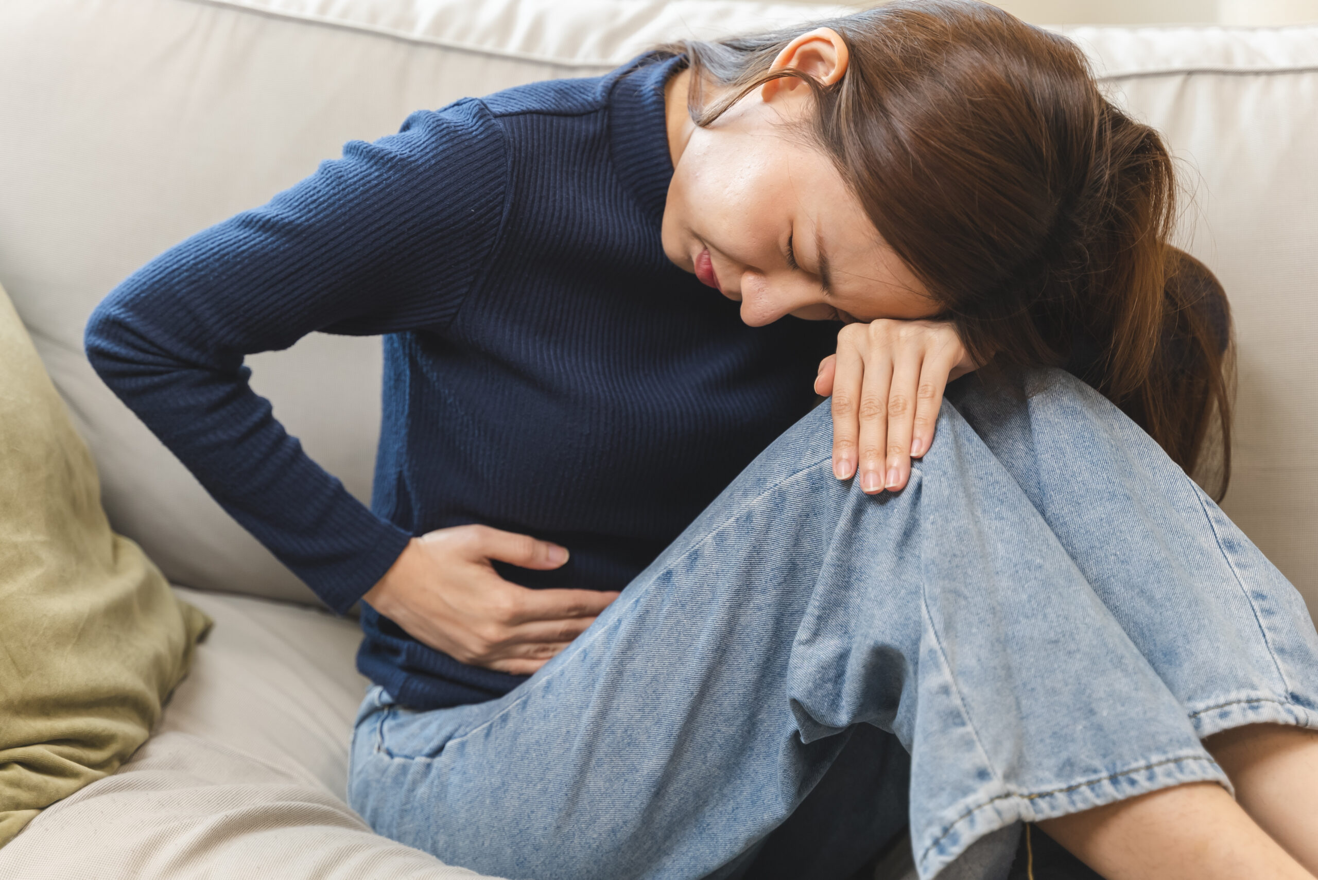 Crampes menstruelles (dysménorrhée) : Symptômes, Causes et Traitements Image d’une femme en pull bleu et jeans, assise sur un canapé, se tenant le ventre en raison de crampes menstruelles, illustrant la douleur liée à la dysménorrhée.