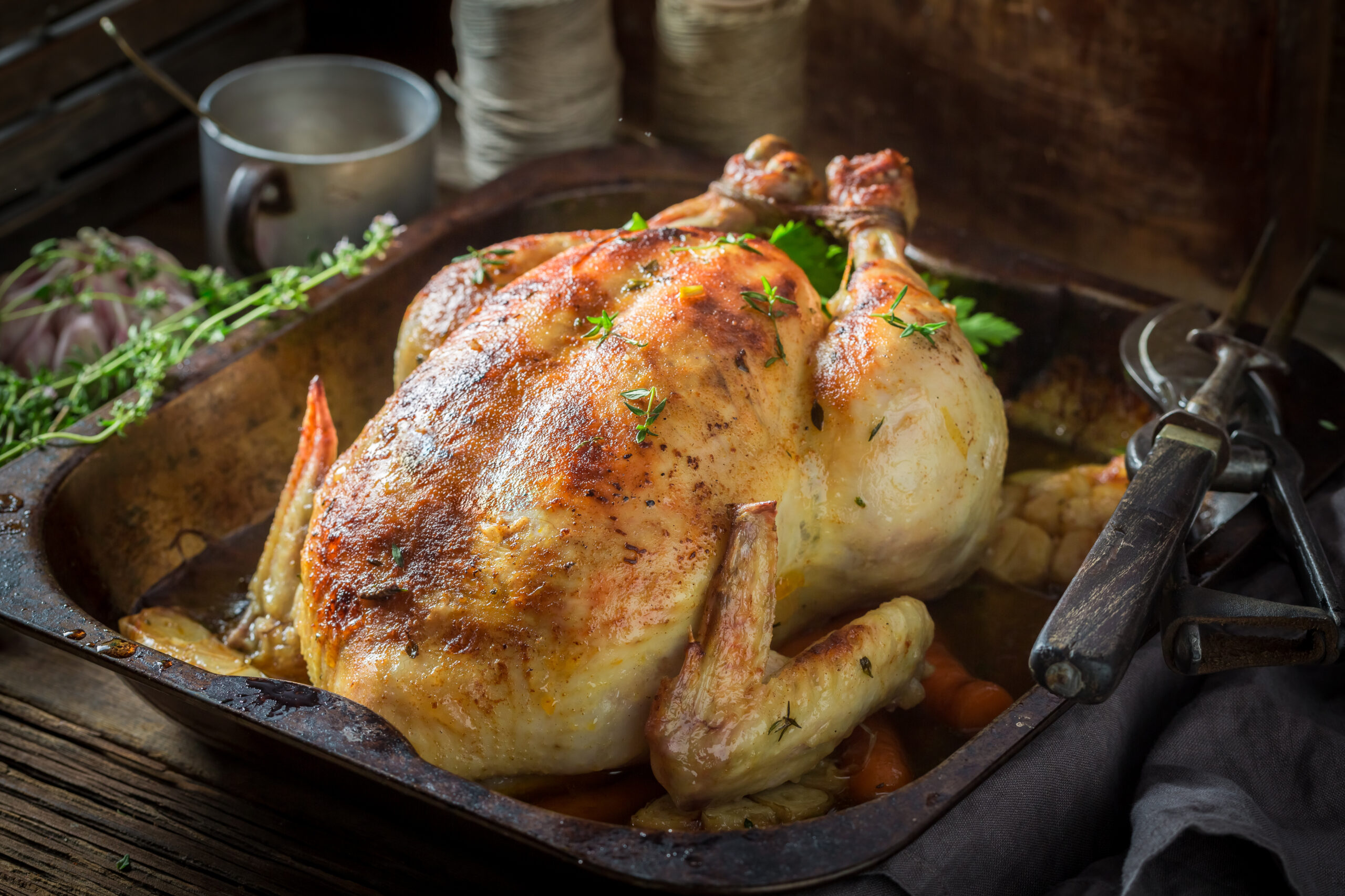 Étude : Manger du poulet pourrait raccourcir votre espérance de vie Poulet rôti aux herbes et légumes dans un plat en fonte, accompagné de thym et d’ail, sur une table en bois avec des ustensiles de cuisine en arrière-plan.