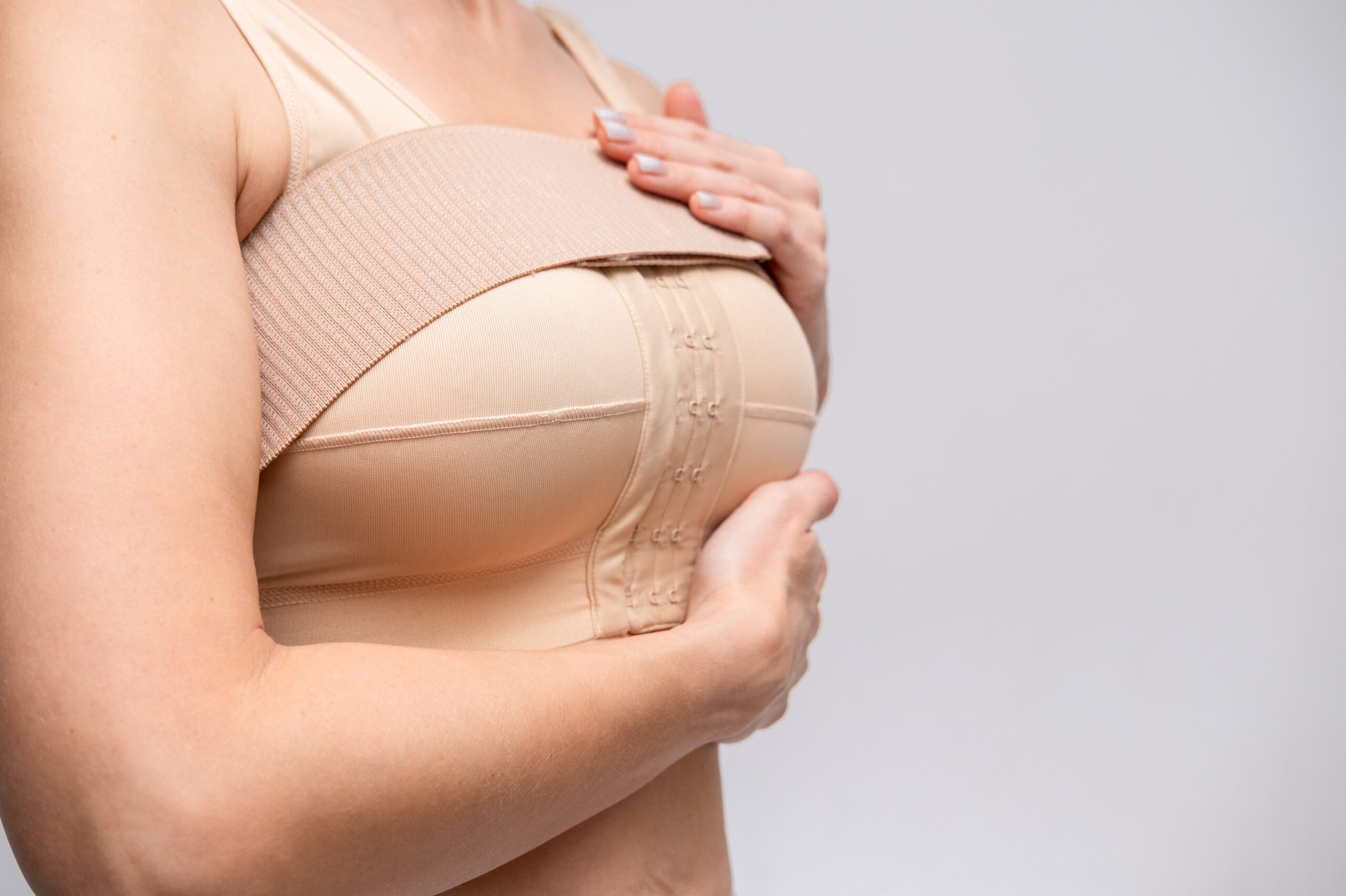 Réduction Mammaire : Tout sur la Chirurgie et ses Bienfaits Une femme portant un soutien-gorge chirurgical beige à fermeture avant, utilisé pour soutenir les seins après une chirurgie de réduction mammaire, sur un fond gris clair.