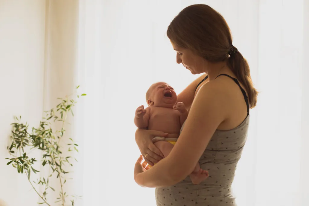Mère tenant son nouveau-né qui pleure dans ses bras, lumière douce en arrière-plan – image représentant les défis émotionnels et physiques du post-partum